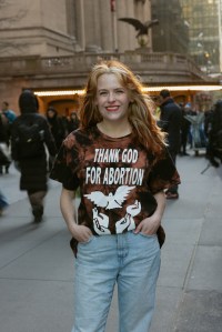 Photo of Becca Rea-Tucker standing on a NYC sidewalk wearing a shirt that reads "Thank God For Abortion" over images of upraised hands and a dove. Becca is smiling at the camera.